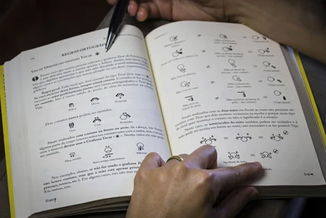 Livro aberto na página "Regras Ortográficas", com uma mão segurando uma caneta