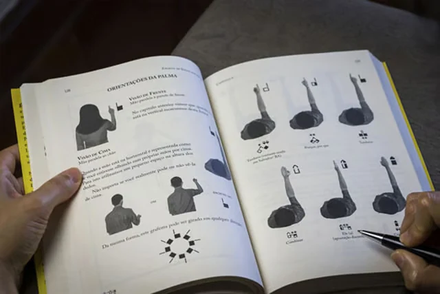 Livro aberto na página "Orientações da Palma", com uma caneta apontando para os grafemas